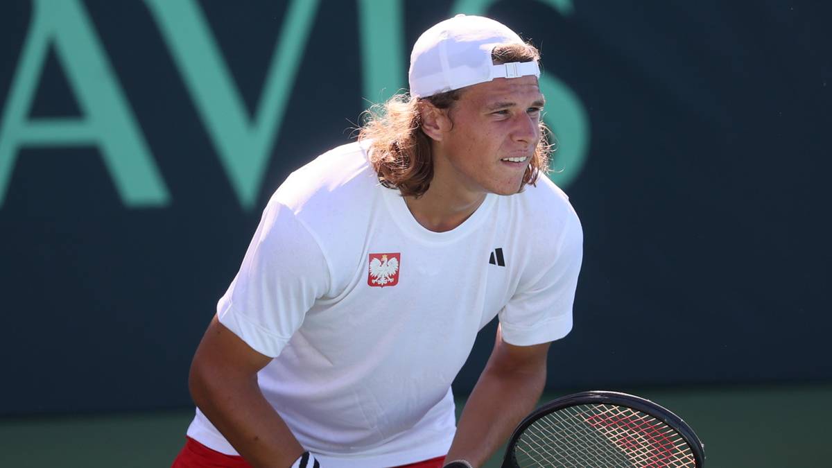 Tomasz Berkieta, tenisista z długimi blond włosami, w białej czapce z daszkiem i białej koszulce z polskim godłem, pochyla się podczas meczu na kortach tenisowych.