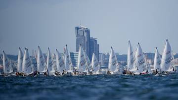 Grupa żagl&oacute;wek ścigających się na wodzie przed miejskim krajobrazem.