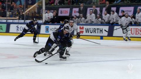 Dw&oacute;ch hokeist&oacute;w w białych strojach stara się odebrać krążek zawodnikowi w ciemnym stroju na lodowisku podczas meczu.