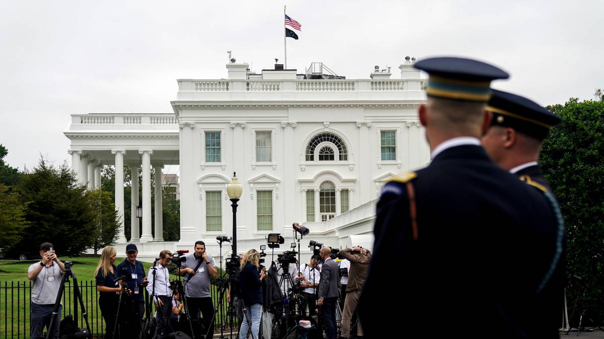 Biały Dom, amerykańska flaga powiewająca na maszcie, żołnierze w mundurach i duża grupa dziennikarzy z aparatami.