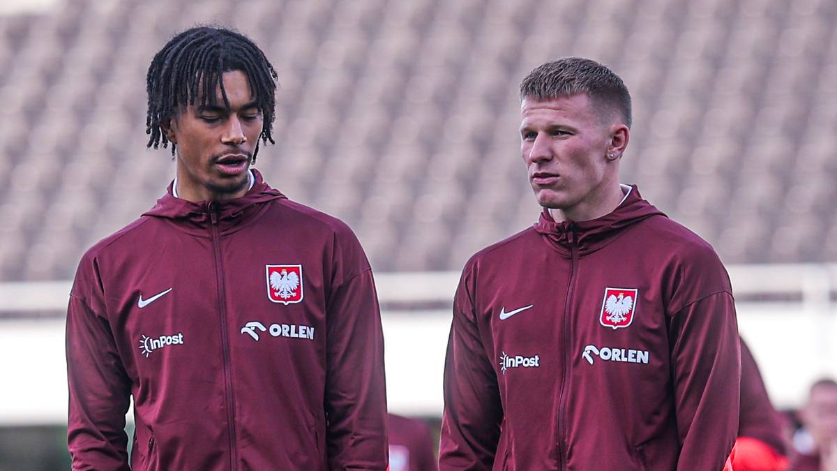 Dw&oacute;ch piłkarzy w bordowych dresach z herbem Polski na piersi stoi na boisku.