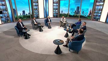Grupa siedmiu os&oacute;b siedzi w okręgu na krzesłach w studiu telewizyjnym. Na środku okręgu znajduje się okrągły dywan, a przed każdym z nich stoi mały stolik z przedmiotami.