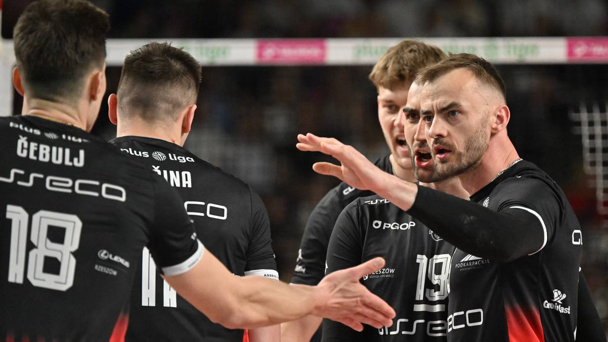Siatkarze w czarnych strojach z czerwonymi elementami, jeden z nich podaje rękę drugiemu.