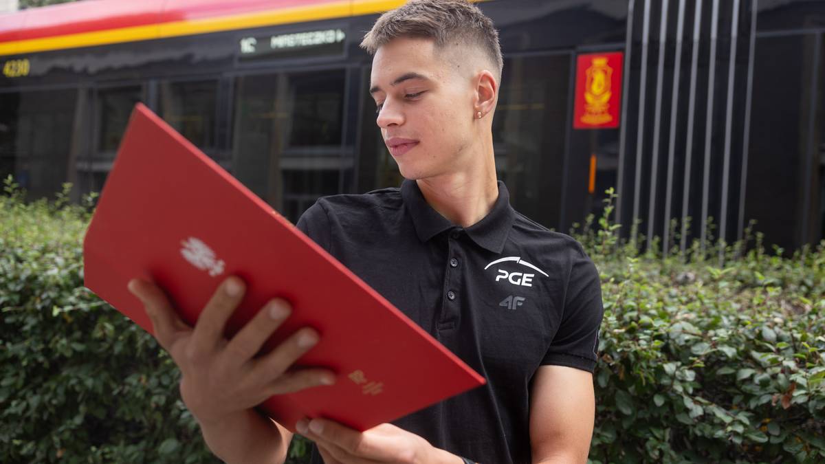 Młody mężczyzna w czarnej koszulce z logo PGE i 4F trzyma przed sobą dużą, czerwoną teczkę. Jest na zewnątrz, za nim widać zielone krzewy i fragment pojazdu komunikacji miejskiej.