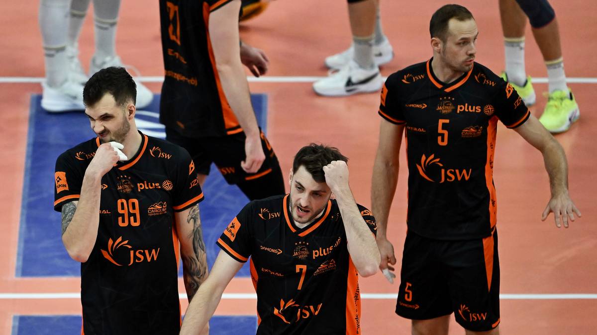 Czterech siatkarzy w czarnych koszulkach z pomarańczowymi wstawkami i numerami na plecach, stojących na parkiecie podczas meczu.