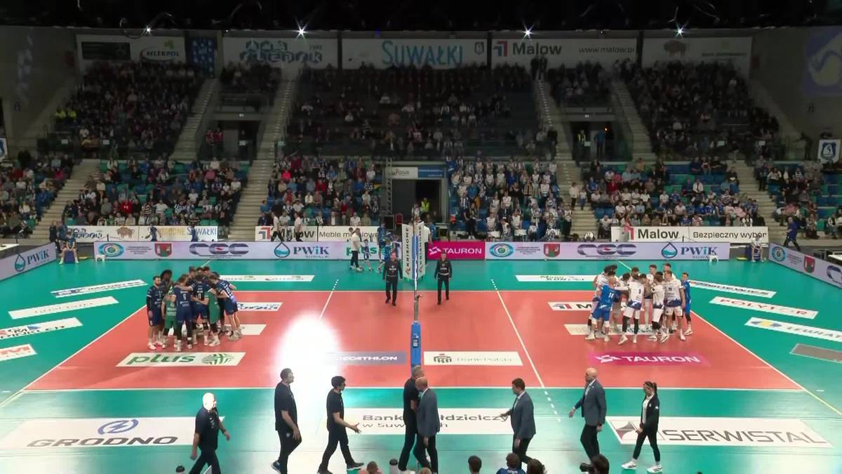 Boisko do siatk&oacute;wki z siatką, na kt&oacute;rym znajdują się dwie drużyny zawodnik&oacute;w w niebieskich i czerwonych strojach. Na trybunach siedzą kibice.