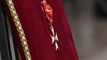 Zbliżenie na ozdobę w kształcie krzyża z białymi ramionami i złotym środkiem, przypiętą do czerwonego materiału z ozdobną złotą plecionką.