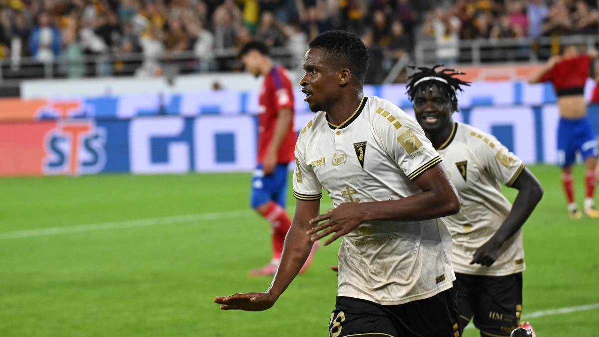 Piłkarz w białej koszulce z złotymi elementami radośnie gestykuluje w stronę kamery. Za nim biegnie inny piłkarz w tej samej koszulce, uśmiechając się. Na drugim planie widać zamazanych piłkarzy w czerwonych strojach oraz fragment stadionu z publicznością i banerami.