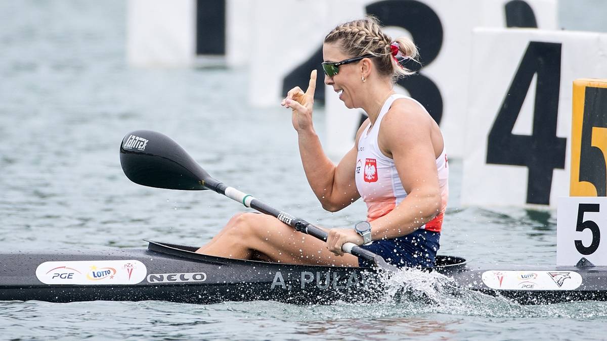Kobieta w biało-czerwonej koszulce siedzi w kajaku i macha palcami na znak zwycięstwa.