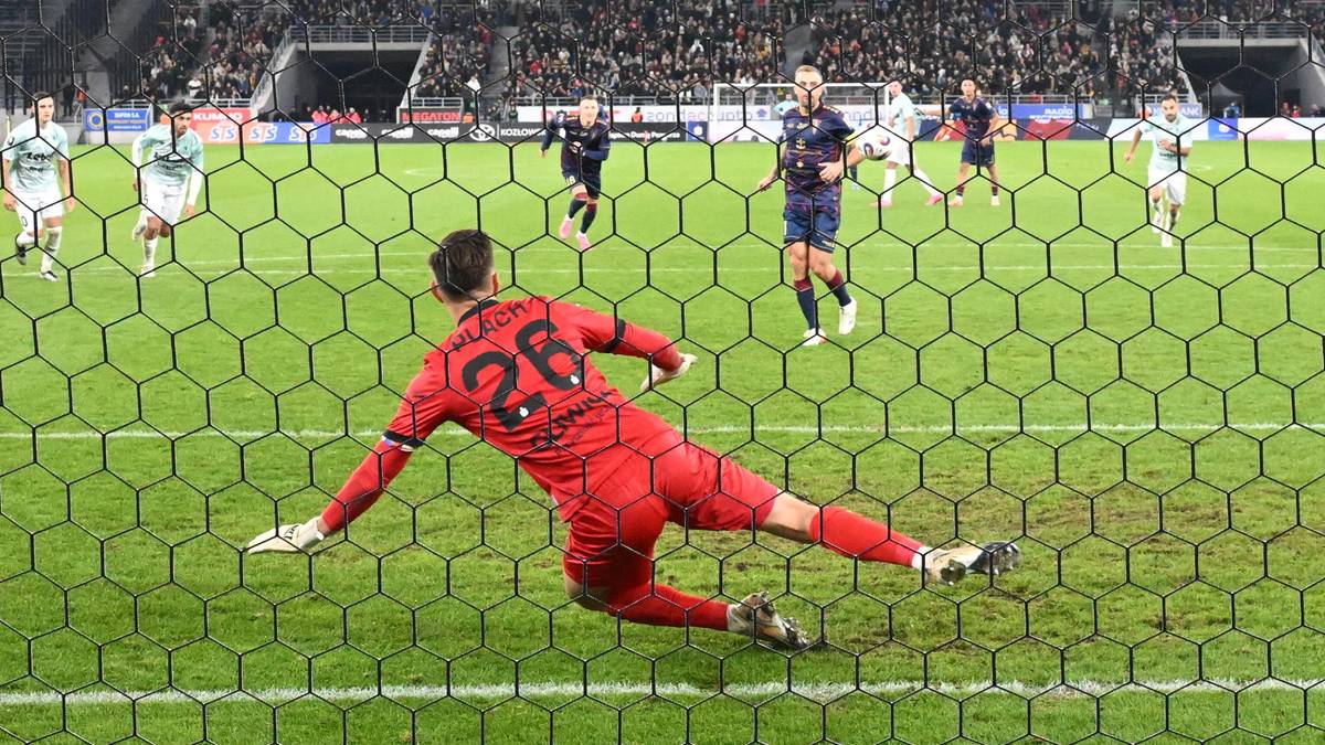 Bramkarz w czerwonym stroju rzuca się w kierunku piłki, próbując ją obronić przed linią bramkową. Na drugim planie widać innych zawodników oraz trybuny stadionu.