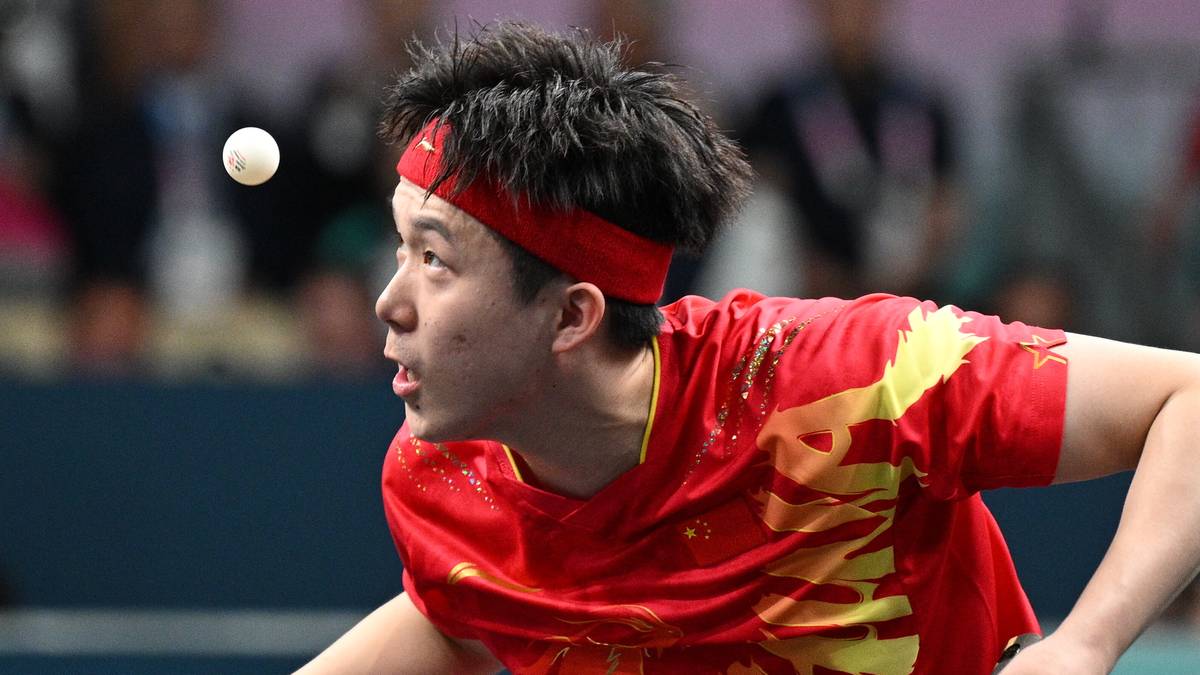 Młody azjatycki zawodnik tenisa stołowego w czerwonej koszulce z ż&oacute;łtymi zdobieniami i czerwonej opasce na czole, przygotowuje się do uderzenia piłeczki.