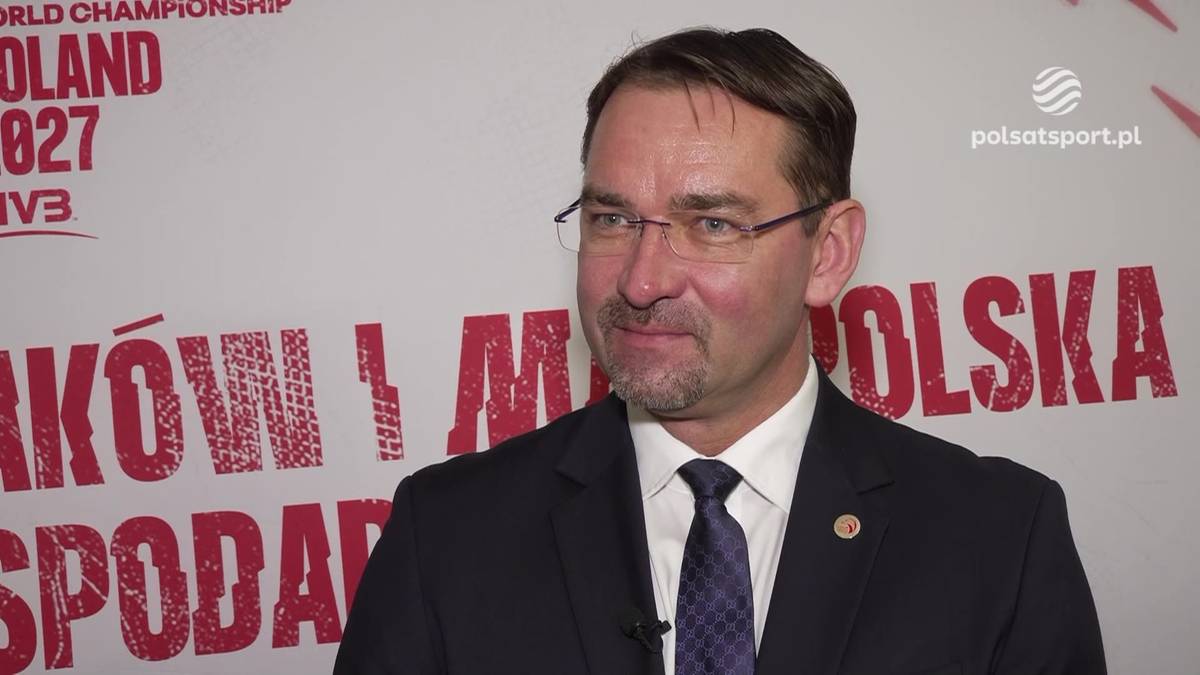 Sebastian Świderski, ubrany w ciemny garnitur i krawat, stoi na tle ściany z napisem "POLSKA". Ma na sobie okulary, a jego twarz wyraża uśmiech.