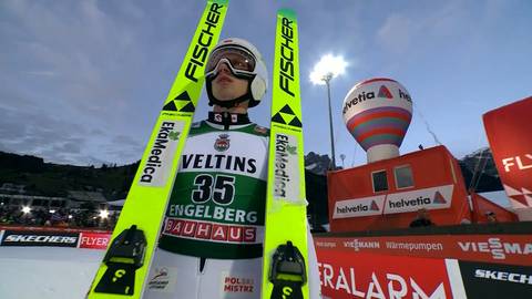 Skoczek narciarski w kasku i goglach stoi z nartami w rękach przed budynkiem z logo "Helvetia" i "Velvetins".