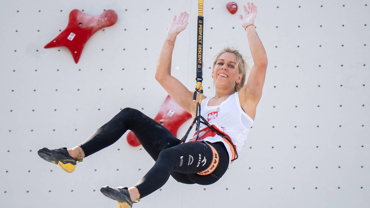 Aleksandra Mirosław podczas zawodów we wspinaczce sportowej, z nogami w powietrzu, uśmiecha się do obiektywu.