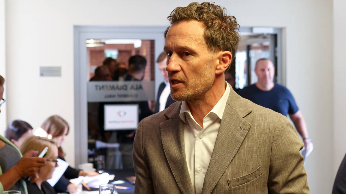 Mężczyzna w jasnej marynarce patrzy w bok, jakby do kogoś mówił. W tle widać inne osoby.