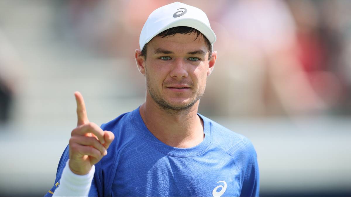 Uśmiechnięty mężczyzna w czapce z daszkiem i niebieskiej koszulce sportowej, wskazujący palcem do góry.