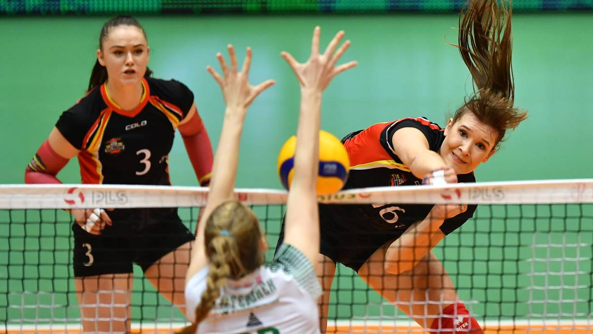 Trzy siatkarki podczas akcji meczowej, dwie po jednej stronie siatki próbują zablokować atak, jedna zawodniczka po drugiej stronie siatki.