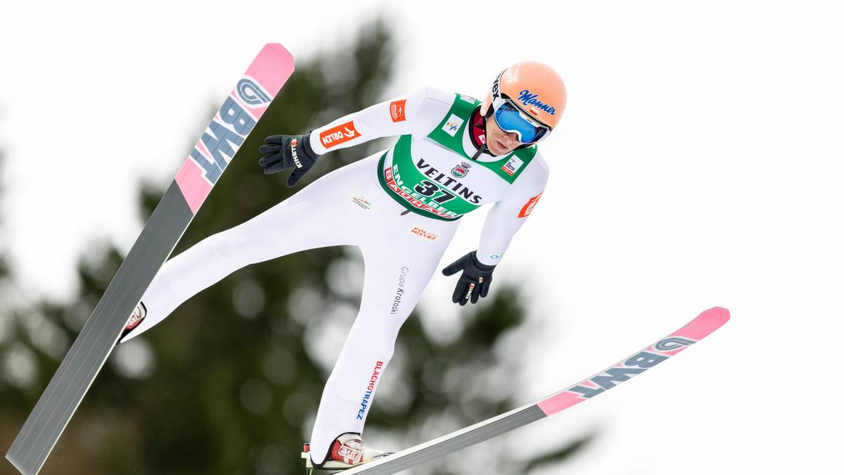 Skoczek narciarski w białym kombinezonie i pomarańczowym kasku wykonuje skok.