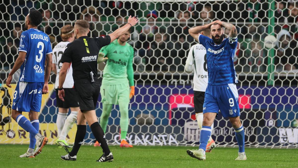 Zawodnicy na boisku piłkarskim, jeden z nich w niebieskim stroju trzyma się za głowę, sędzia w czarnym stroju pokazuje ręką.