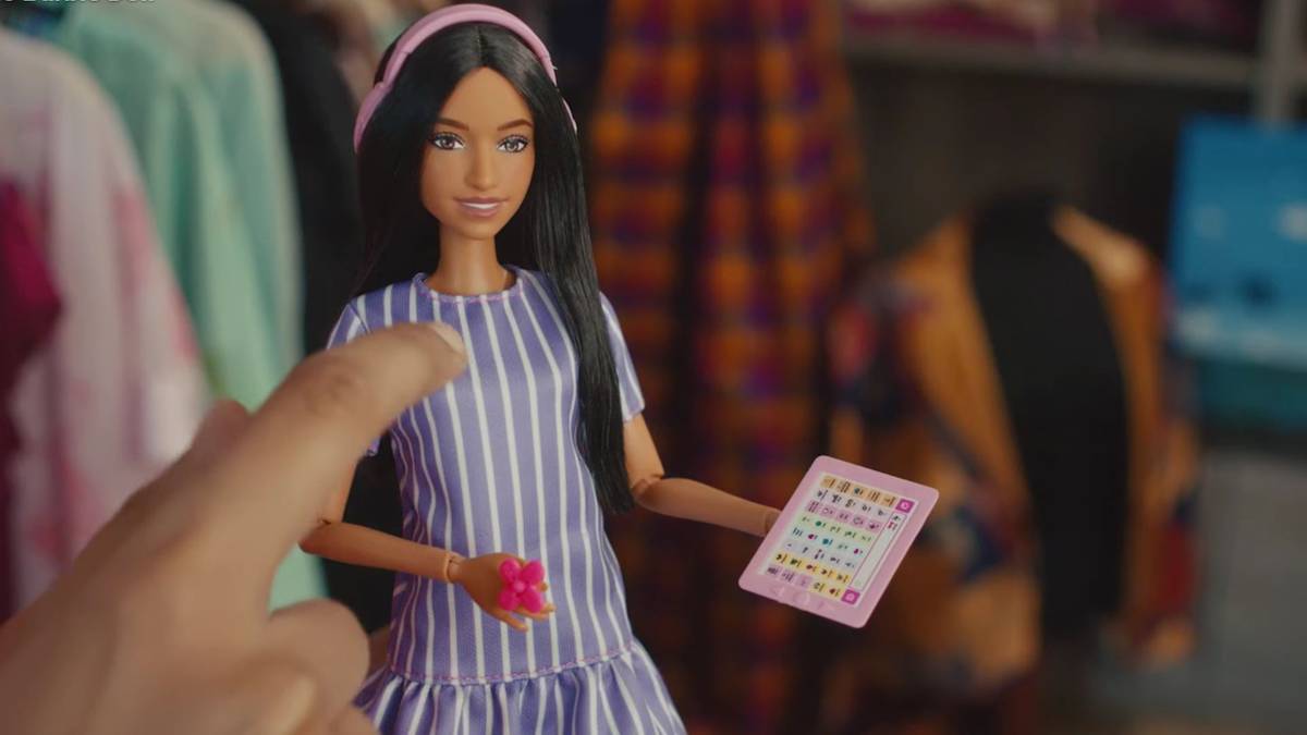 Lalka Barbie w niebiesko-białej sukience w paski, trzymająca w dłoni r&oacute;żową bransoletkę i r&oacute;żową zabawkę w kształcie serca. Lalka stoi przed regałem z ubraniami.