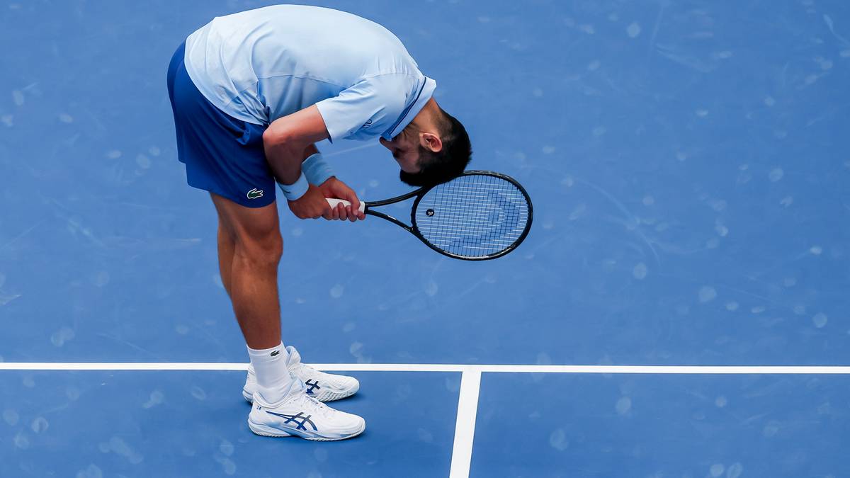 Tenisista Novak Djoković pochyla się nad niebieskim kortem z rakietą w ręku, z głową opuszczoną w geście frustracji lub rozpaczy.