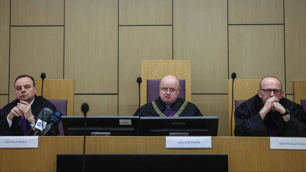 Trzech sędzi&oacute;w w togach sądowych siedzi za stołem w sali sądowej przed herbem Polski.