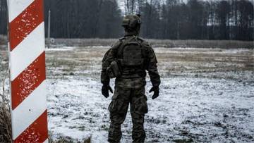 Żołnierz w pełnym umundurowaniu stoi tyłem do widza przy słupie granicznym, na tle zimowego krajobrazu.