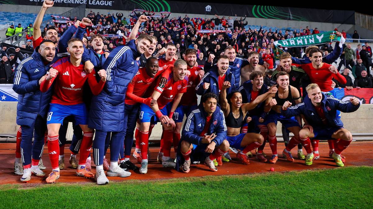 Drużyna piłkarska w czerwono-niebieskich strojach pozuje do zdjęcia na boisku, świętując zwycięstwo przed trybunami pełnymi kibic&oacute;w.