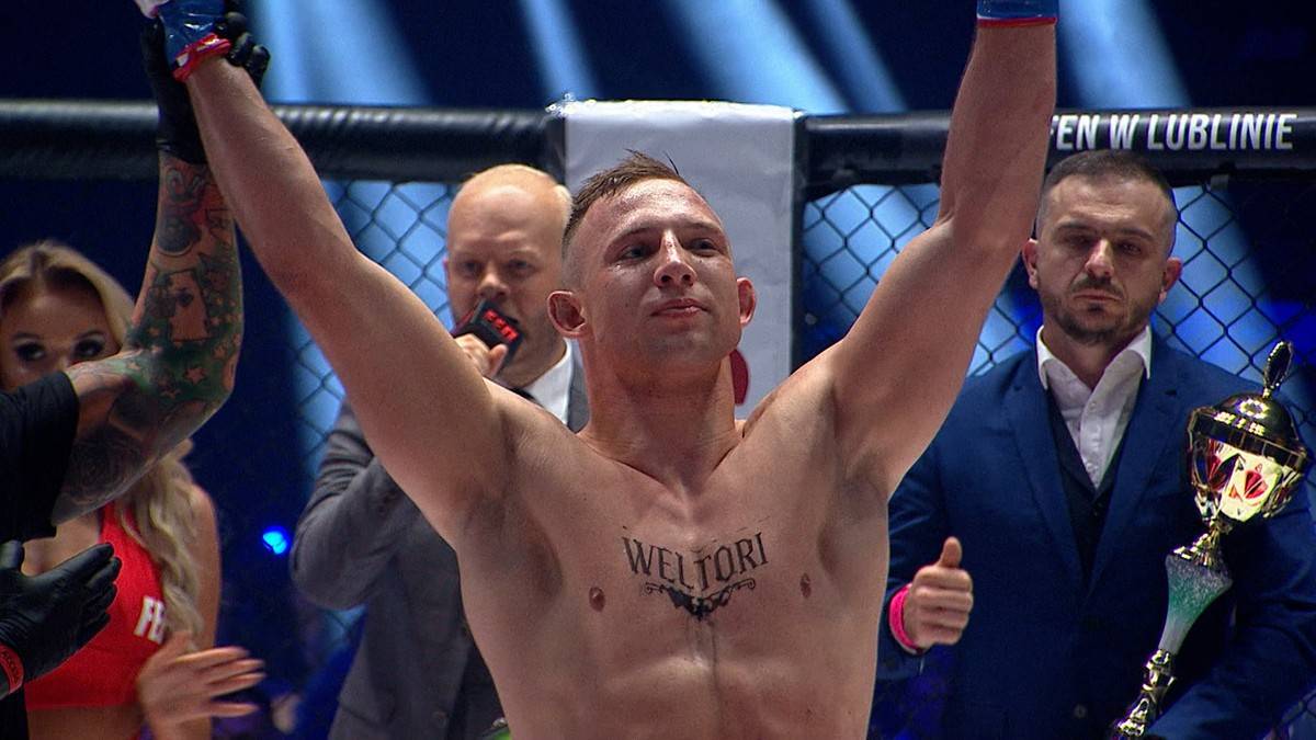 Zawodnik MMA z uniesionymi rękami w geście zwycięstwa, z tatuażem na klatce piersiowej i trofeum w dłoni.