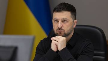 Wołodymyr Zełenski, prezydent Ukrainy, siedzi przy stole z rękami złożonymi przed brodą, na tle flagi Ukrainy.