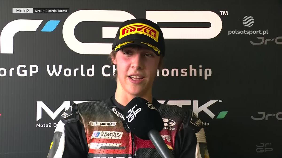 Młody zawodnik motocyklowy w czapce z daszkiem i stroju wyścigowym, z mikrofonem przed twarzą, podczas wywiadu po zawodach.