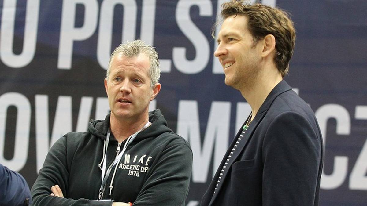 Dwóch mężczyzn, z lewej w czarnej bluzie z kapturem z napisem "NIKE", z prawej w granatowym garniturze, patrzą w bok. W tle białe napisy na ciemnym tle.