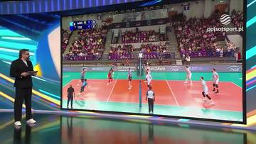 Duży ekran telewizyjny wyświetlający transmisję meczu siatk&oacute;wki. Na boisku widać zawodnik&oacute;w w białych i czerwonych strojach, a na trybunach kibic&oacute;w. Obok ekranu stoi mężczyzna w czarnym garniturze, patrzący na ekran i trzymający w ręku tablet.