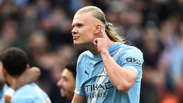 Piłkarz Erling Haaland w jasnoniebieskiej koszulce Manchesteru City z numerem 9 na plecach, odwr&oacute;cony w prawo, z ręką na szyi, na tle innych piłkarzy i publiczności.