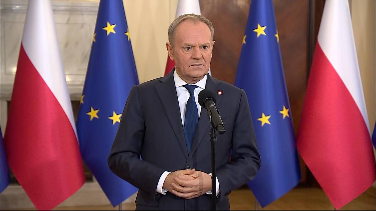 Premier Donald Tusk przemawia na konferencji prasowej, na tle flag Polski i Unii Europejskiej.