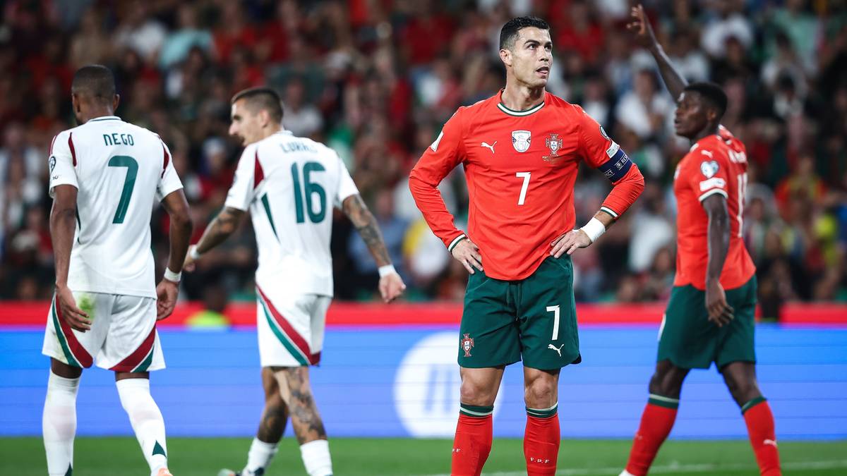 Cristiano Ronaldo w czerwonym stroju reprezentacji Portugalii z numerem 7, stojący na boisku piłkarskim podczas meczu. Obok niego inni zawodnicy w białych strojach.