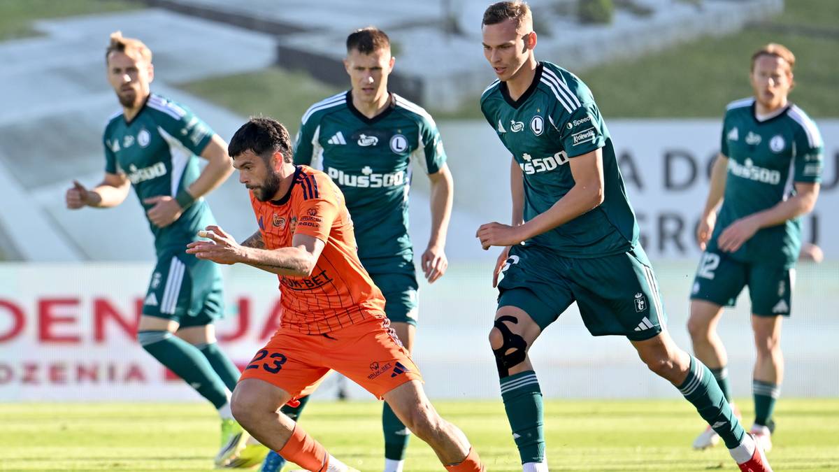 Piłkarz w pomarańczowym stroju walczy o piłkę z zawodnikiem w zielonym stroju.