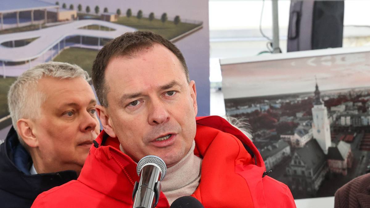 Mężczyzna w czerwonej bluzie przemawia do mikrofonu, w tle inny mężczyzna