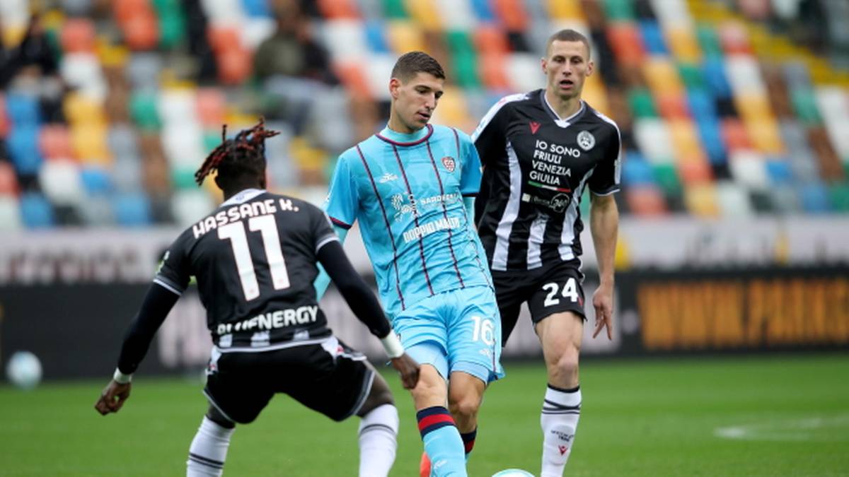 Trzech piłkarzy w akcji na boisku piłkarskim podczas meczu. Jeden z nich, w niebieskim stroju, kontroluje piłkę. Dwóch przeciwników w czarnych koszulkach z białymi pasami próbuje mu przeszkodzić.