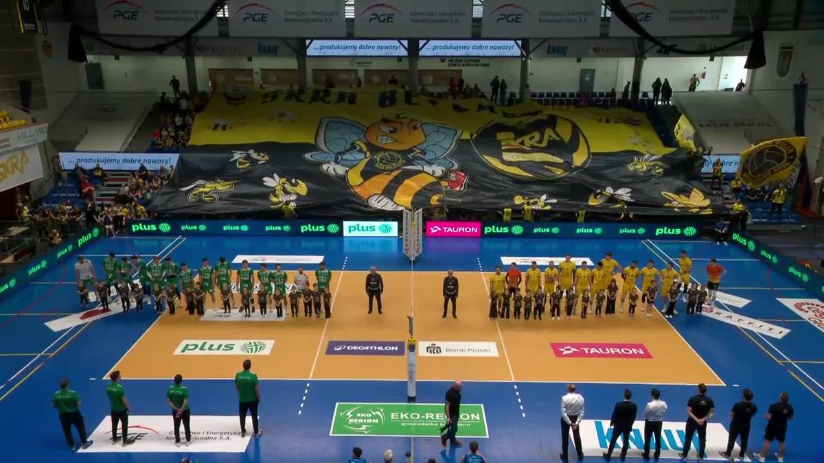 Drużyny siatkarskie stoją po przeciwnych stronach boiska przed rozpoczęciem meczu, z widownią w tle i transparentem.