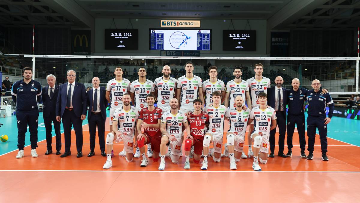 Drużyna siatkarska w białych i czerwonych strojach pozuje do zdjęcia na tle siatki i trybun.