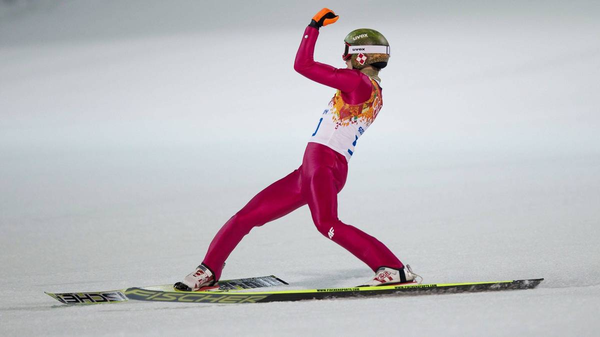 Skoczek narciarski w r&oacute;żowym kombinezonie i z zielonym kaskiem triumfalnie unosi rękę po skoku.