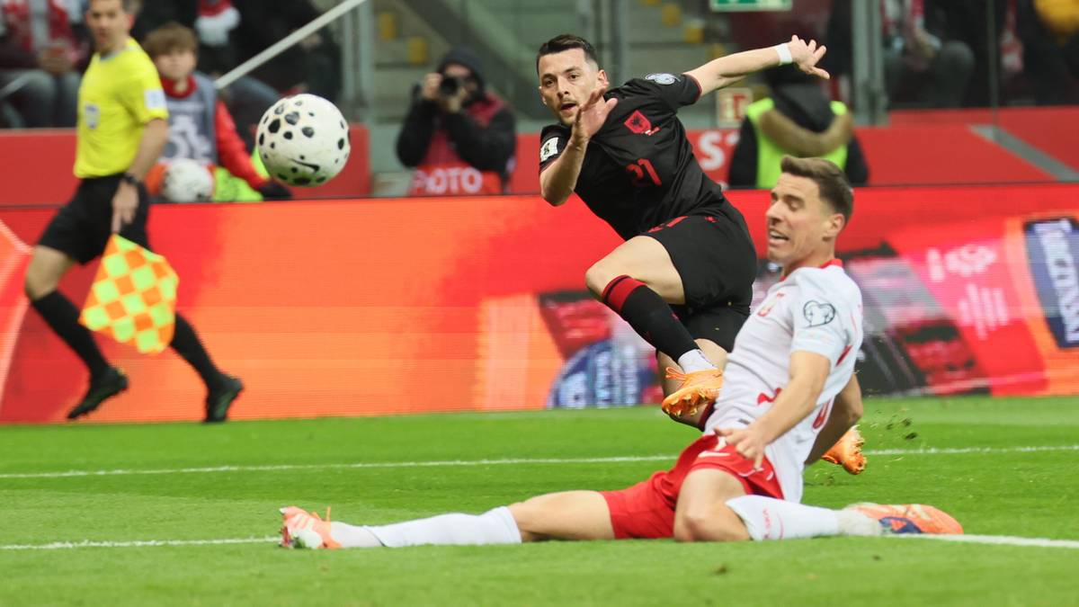 Piłkarz w białej koszulce leży na murawie, a obok niego drugi zawodnik w czarnej próbuje odebrać mu piłkę.