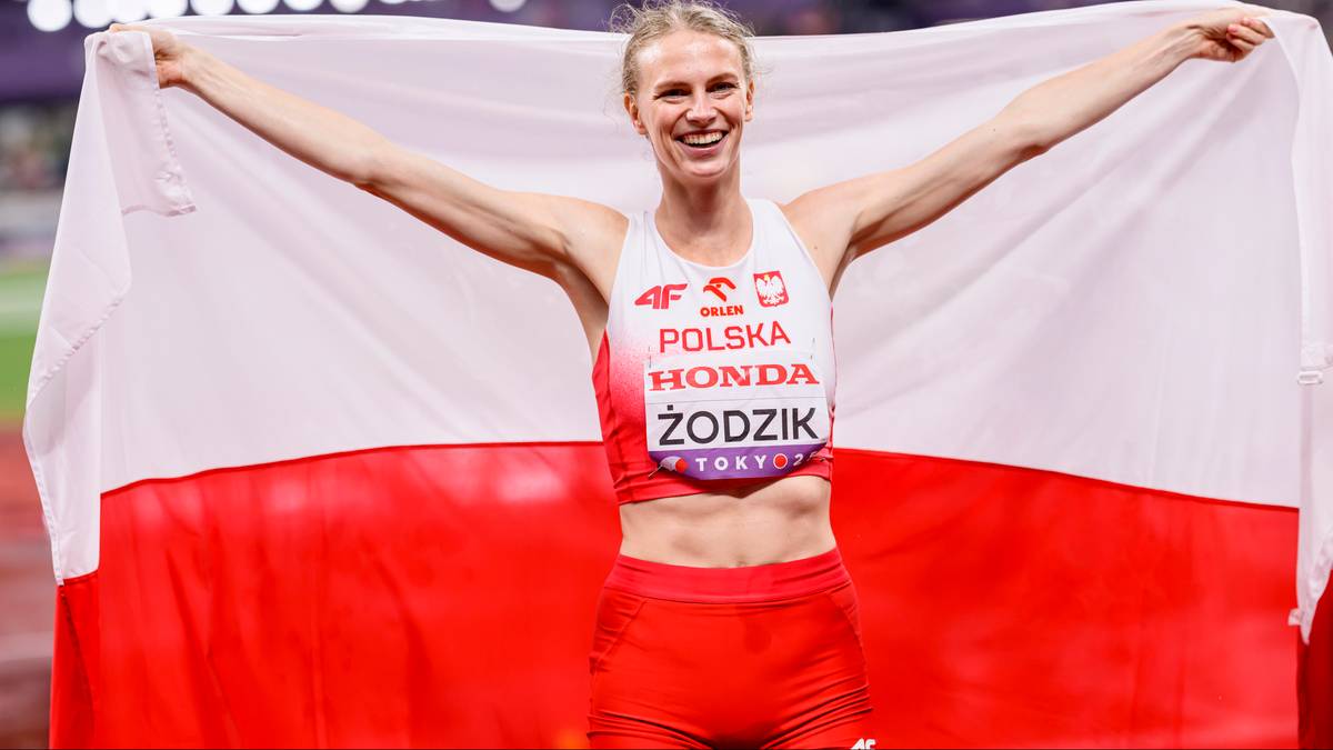 Uśmiechnięta lekkoatletka z flagą Polski rozwiniętą za plecami, patrząca w obiektyw.