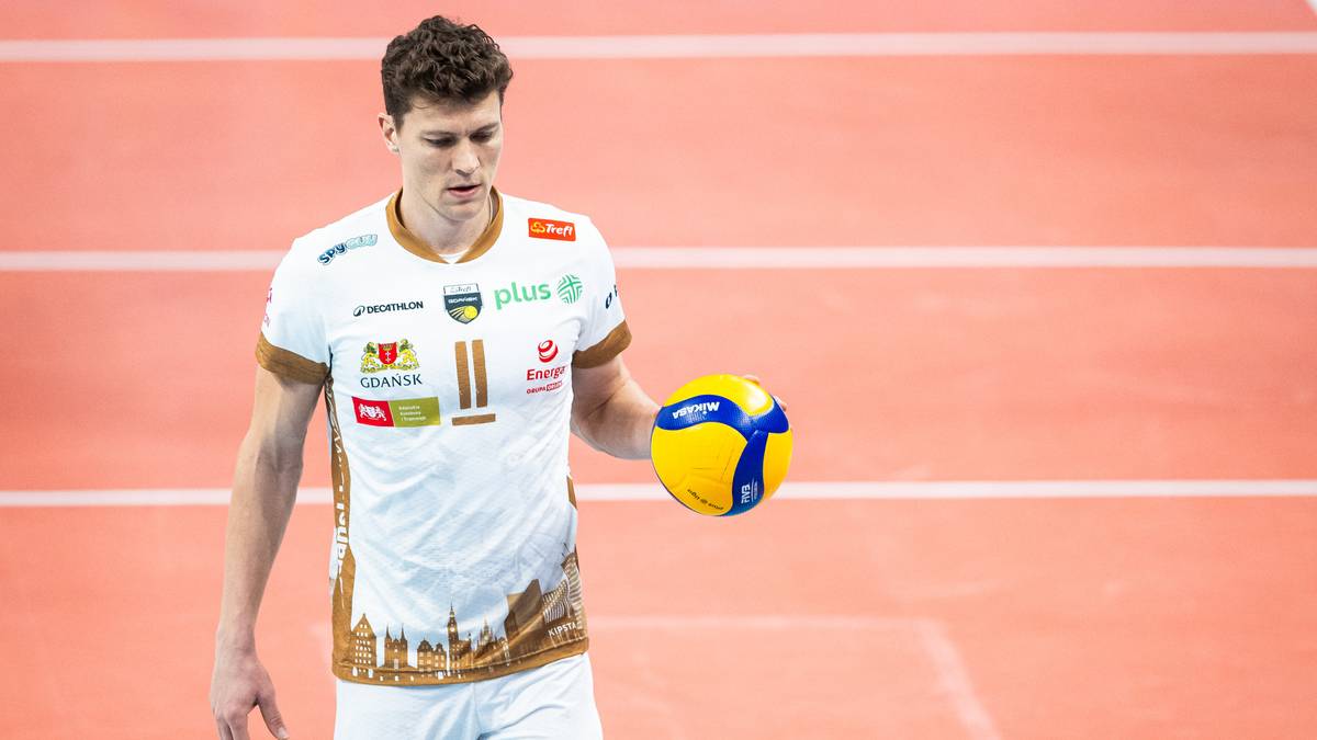 Młody siatkarz w białej koszulce z logo PlusLiga, trzymający w dłoni ż&oacute;łto-niebieską piłkę siatkową. Jest na boisku z pomarańczową nawierzchnią i białymi liniami.