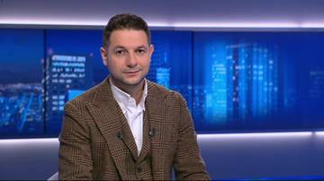 Patryk Jaki siedzi w studiu telewizyjnym, ma na sobie brązową marynarkę w kratę i białą koszulę. W tle widać panel z ekranami wyświetlającymi nocną panoramę miasta.