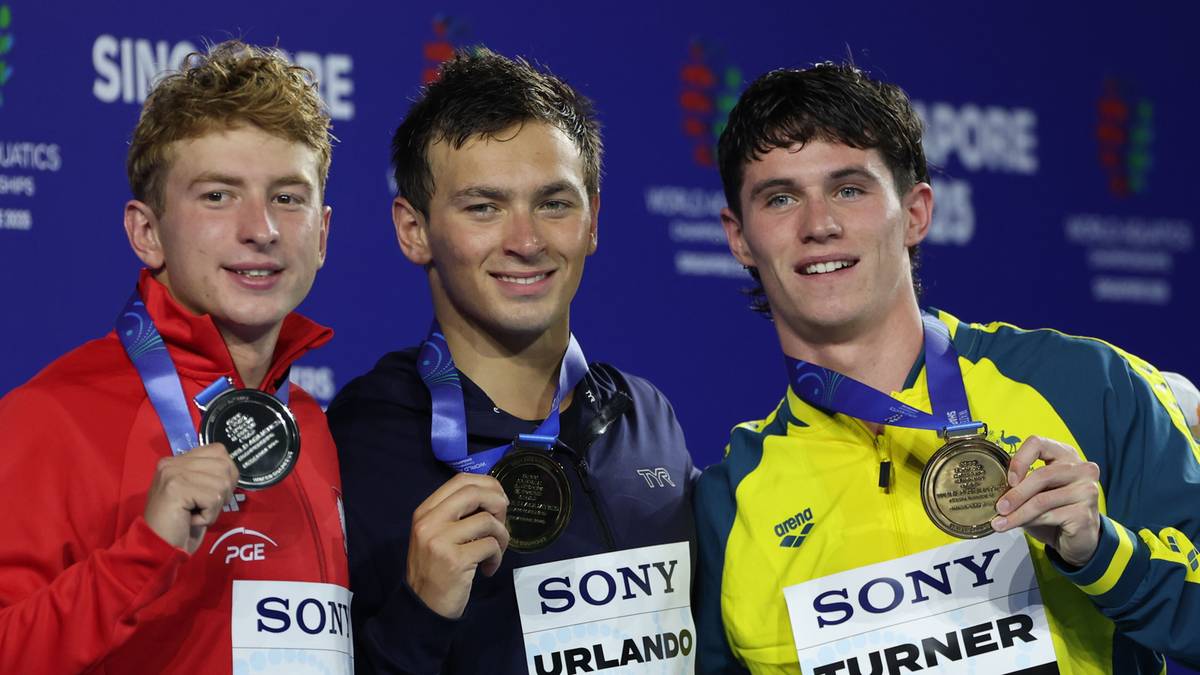 Trzech młodych mężczyzn pozuje do zdjęcia z medalami na szyjach. Wszyscy są uśmiechnięci. Jeden z nich, w czerwonym swetrze, trzyma medal w ręku. Dwóch pozostałych ma medale na szyjach, a jeden z nich pokazuje go do kamery.