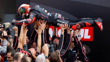 Motocyklista w czarno-czerwonym kombinezonie z zielonymi elementami na kasku jest unoszony przez tłum ludzi.