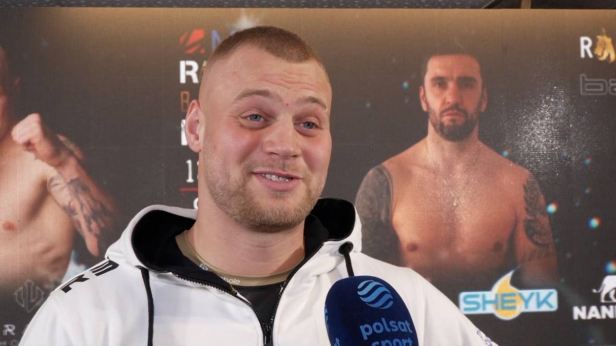 Uśmiechnięty mężczyzna w bluzie z kapturem, stojący przed plakatem z innymi bokserami.