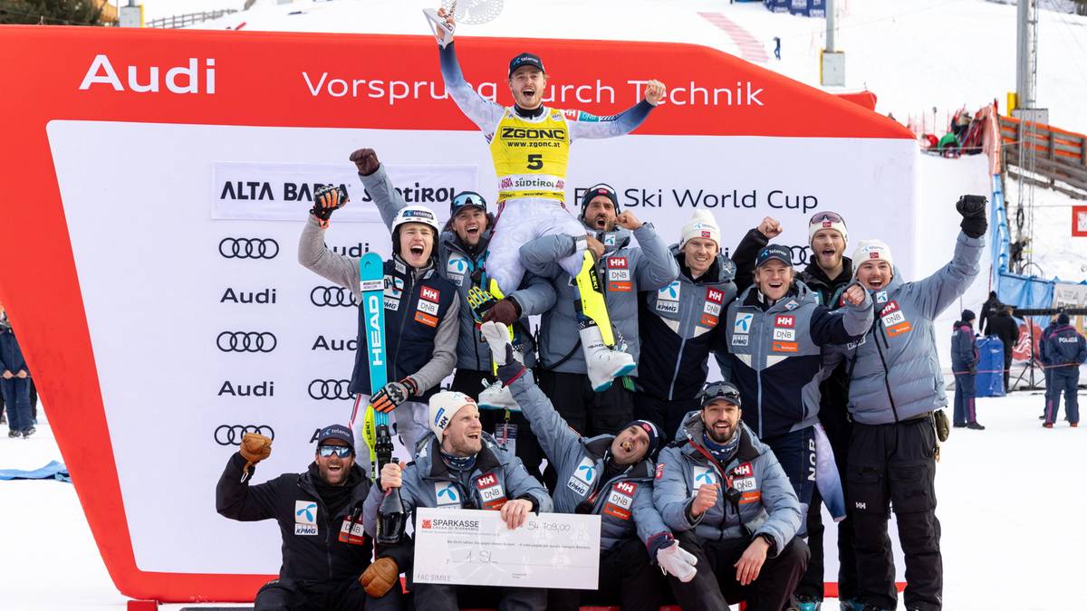 Grupa narciarzy na podium z czekiem i pucharami, w tle tablica wynik&oacute;w zawod&oacute;w Audi Ski World Cup.